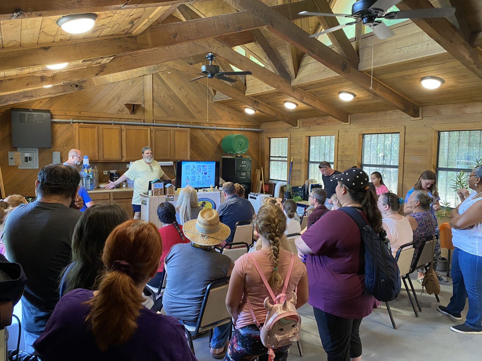 Residents gathered at a community meeting inside a wood-paneled pavilion, listening to a presentation